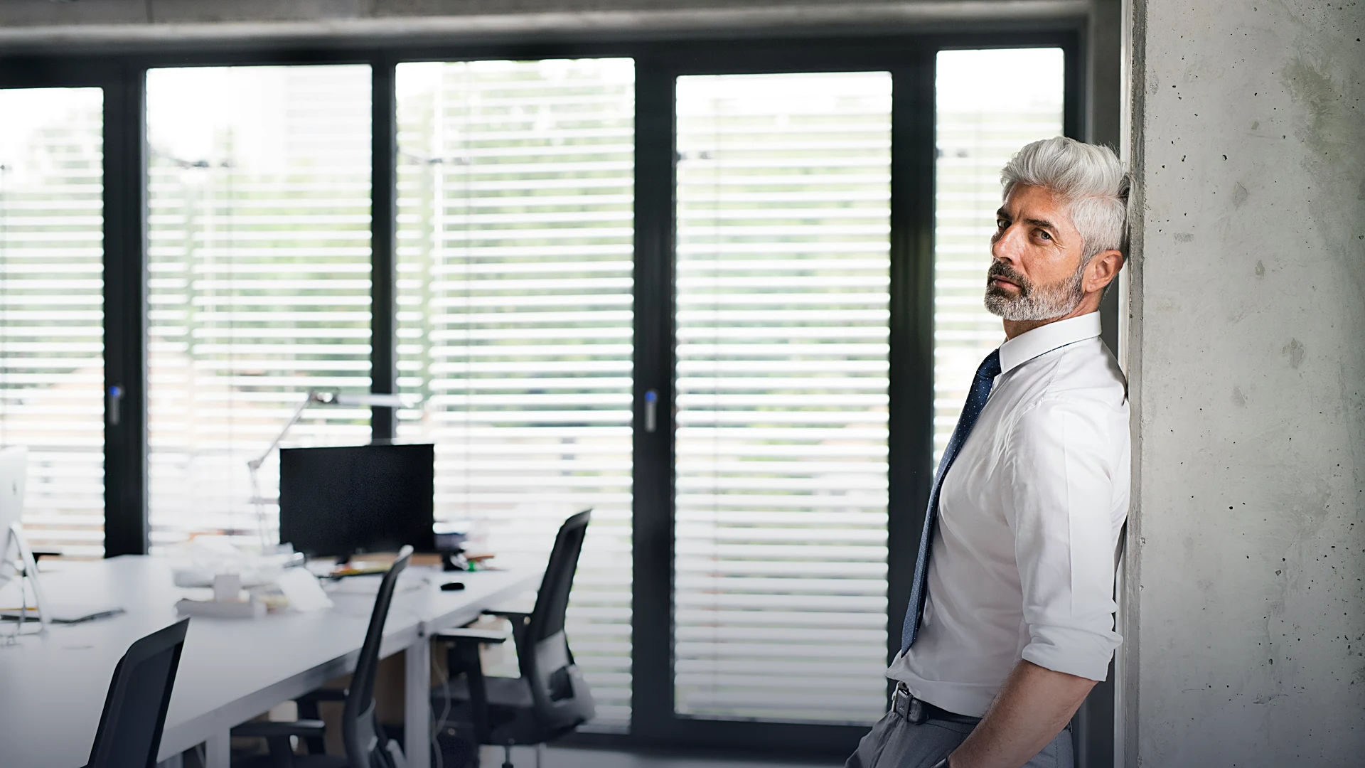 A leader leaning against a pillar beside an office of empty desks and vacant chairs — the disruption already underway inside the organization